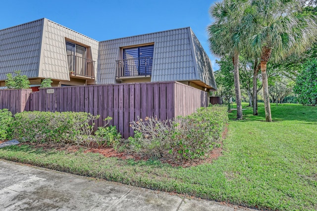 view of side of property featuring mansard roof, a lawn, a balcony, a tile roof, and fence