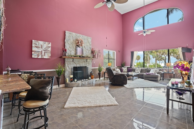 tiled living room with ceiling fan, a towering ceiling, and a stone fireplace