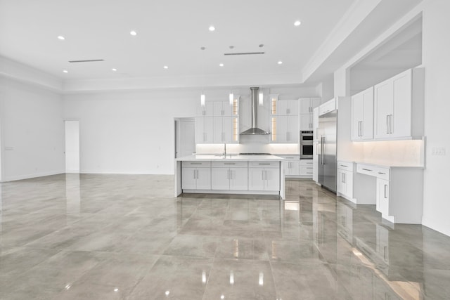 kitchen featuring decorative backsplash, a raised ceiling, wall chimney exhaust hood, a kitchen island with sink, and white cabinetry