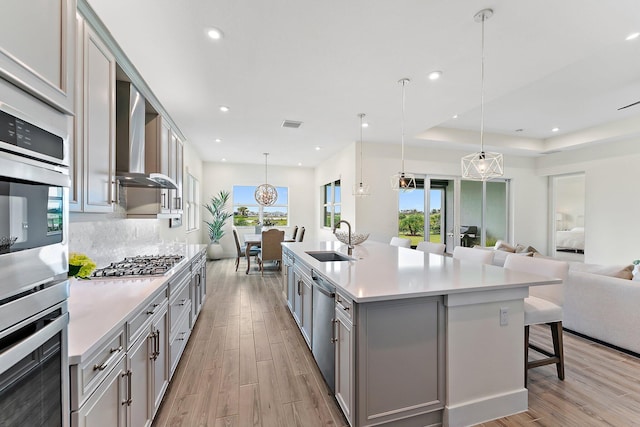 kitchen with wall chimney range hood, light wood-type flooring, an island with sink, and sink