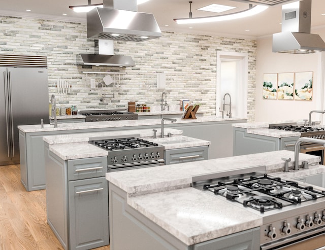 kitchen featuring wall chimney exhaust hood, island exhaust hood, an island with sink, light stone counters, and light wood-type flooring