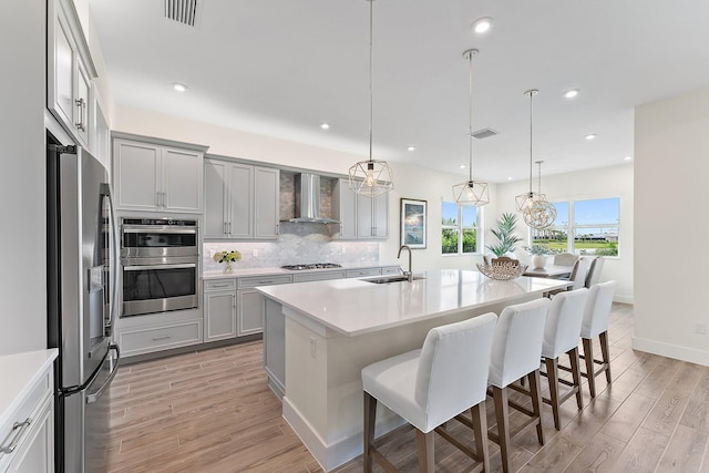 kitchen featuring a kitchen island with sink, a kitchen breakfast bar, wall chimney exhaust hood, appliances with stainless steel finishes, and gray cabinetry