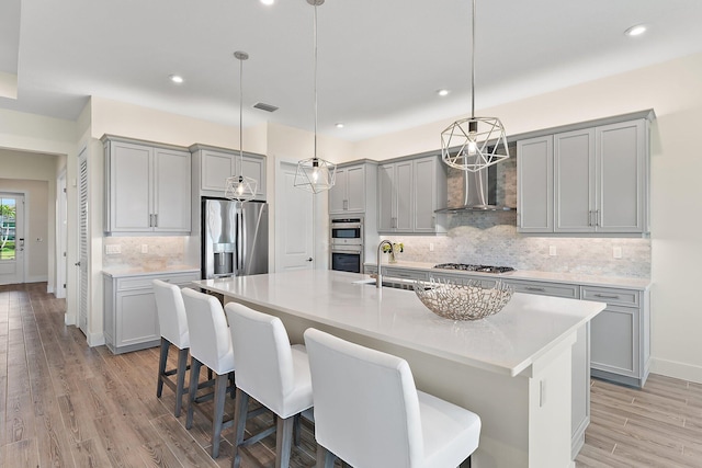 kitchen with a center island with sink, light hardwood / wood-style flooring, appliances with stainless steel finishes, sink, and wall chimney range hood