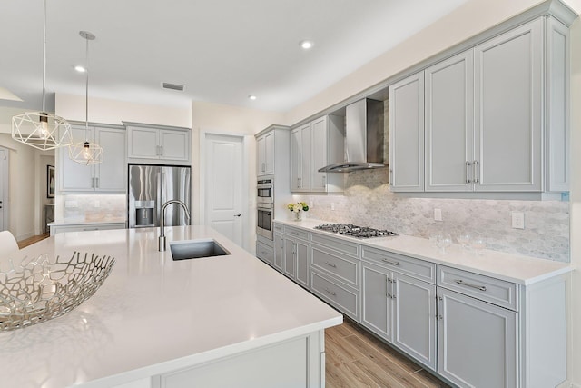 kitchen with light wood-type flooring, appliances with stainless steel finishes, sink, wall chimney range hood, and a center island with sink