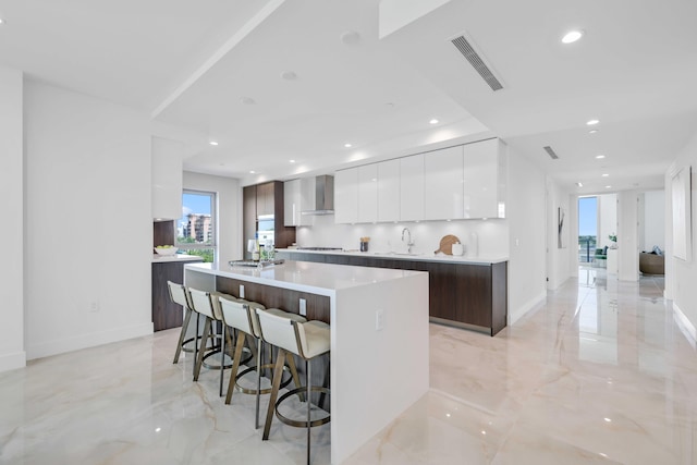 kitchen featuring light tile patterned flooring, dark brown cabinetry, wall chimney exhaust hood, an island with sink, and white cabinets