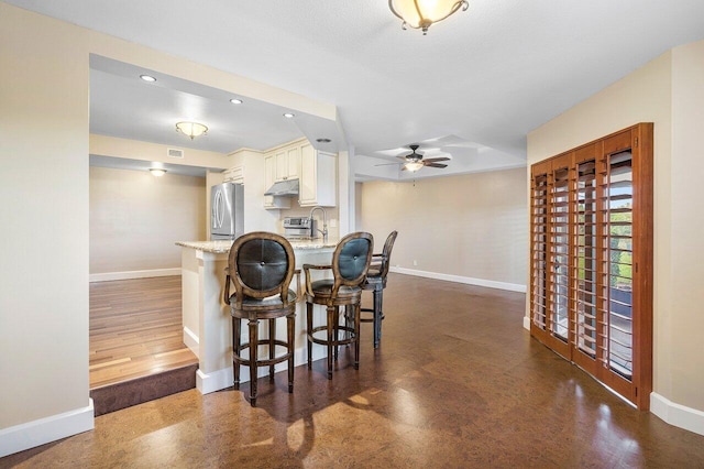 kitchen with under cabinet range hood, a peninsula, a breakfast bar, baseboards, and freestanding refrigerator