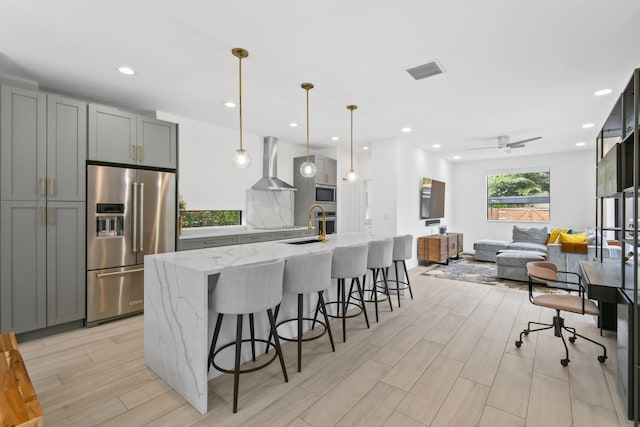 kitchen featuring appliances with stainless steel finishes, light stone countertops, decorative light fixtures, wall chimney range hood, and gray cabinetry