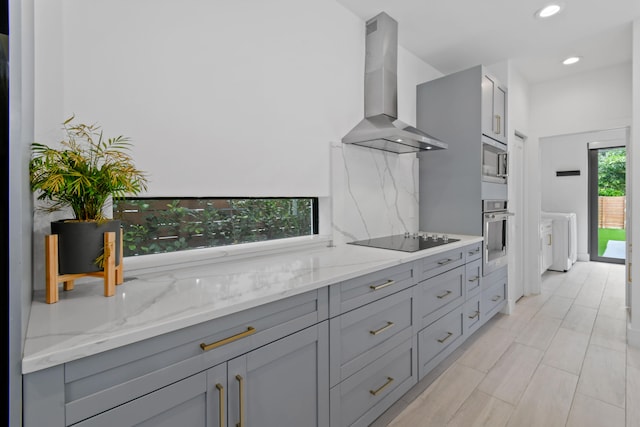 kitchen with gray cabinets, stainless steel appliances, wall chimney range hood, and light stone countertops