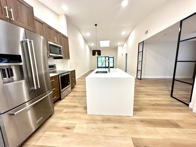 kitchen with light wood-type flooring, a kitchen island with sink, sink, decorative backsplash, and appliances with stainless steel finishes