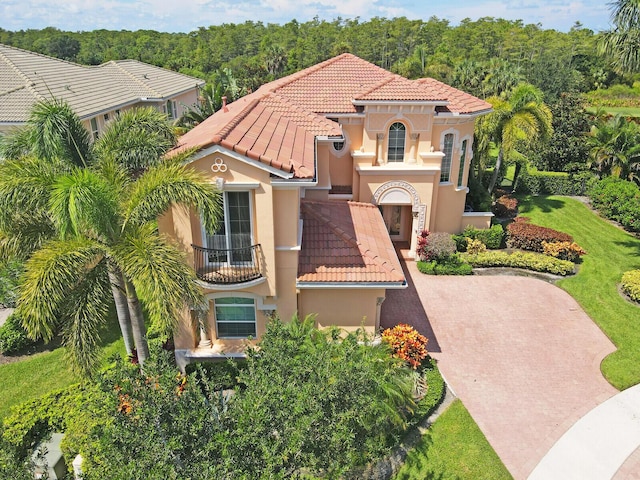 mediterranean / spanish-style home with a balcony, a forest view, a tile roof, decorative driveway, and stucco siding