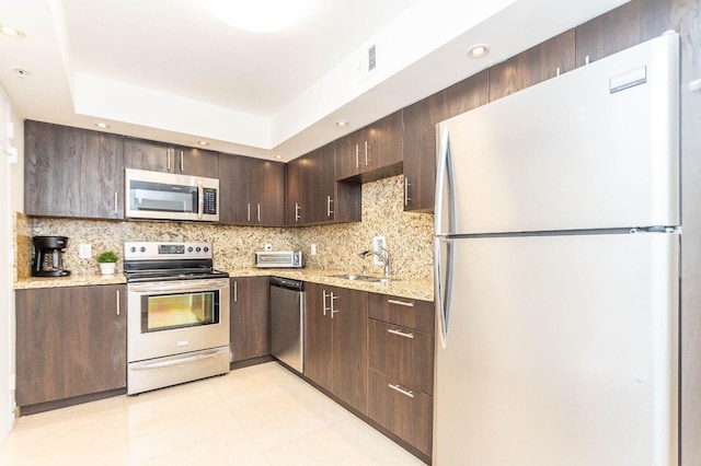 kitchen with stainless steel appliances, light stone counters, sink, a raised ceiling, and dark brown cabinetry