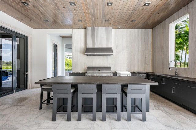 kitchen with wall chimney range hood, wood ceiling, a kitchen island, sink, and light tile patterned flooring