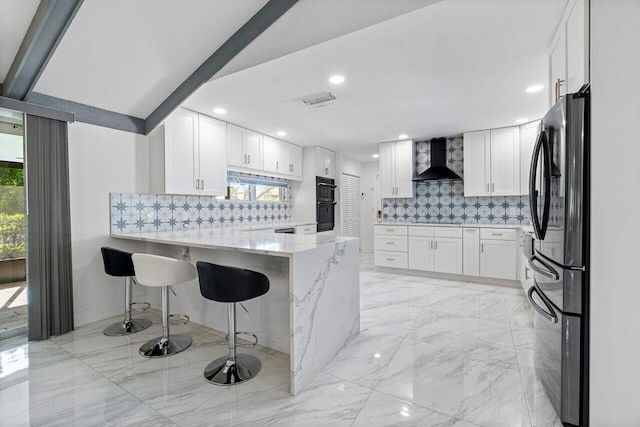 kitchen with white cabinets, kitchen peninsula, wall chimney range hood, a kitchen breakfast bar, and refrigerator