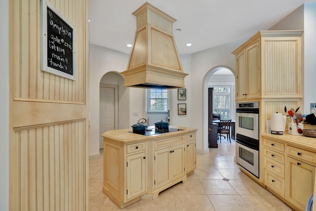 kitchen with black electric stovetop, double oven, light brown cabinetry, light tile patterned flooring, and custom exhaust hood