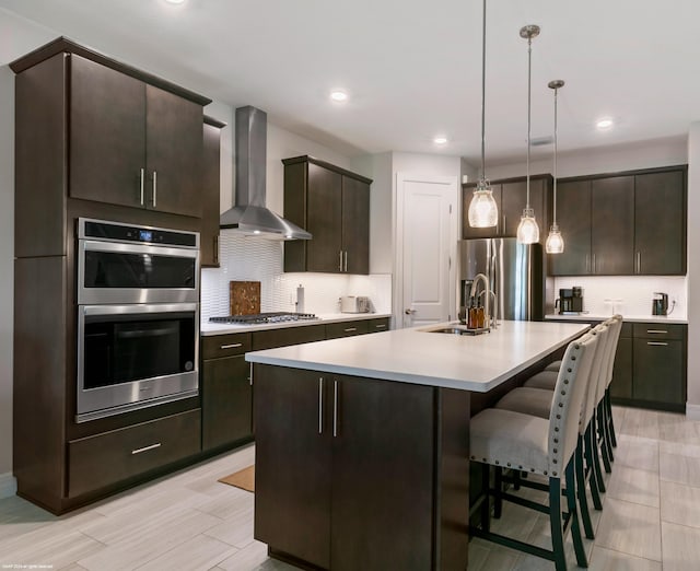 kitchen with stainless steel appliances, dark brown cabinets, light countertops, wall chimney range hood, and a sink