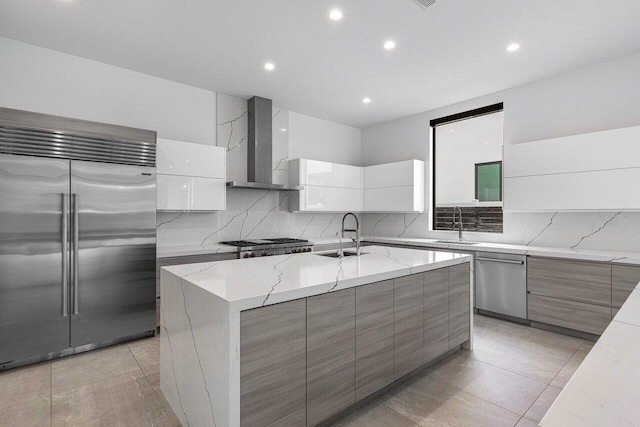 kitchen featuring wall chimney range hood, an island with sink, appliances with stainless steel finishes, white cabinetry, and light stone countertops