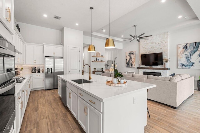 kitchen featuring a stone fireplace, an island with sink, stainless steel appliances, sink, and ceiling fan