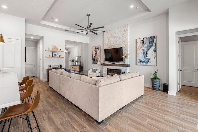 living room featuring light wood-type flooring, ceiling fan, a raised ceiling, and a stone fireplace