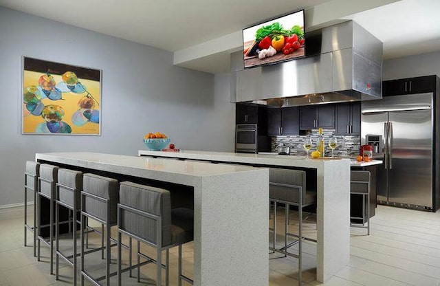 interior space with wall chimney range hood, a kitchen island, tasteful backsplash, a kitchen bar, and stainless steel built in fridge