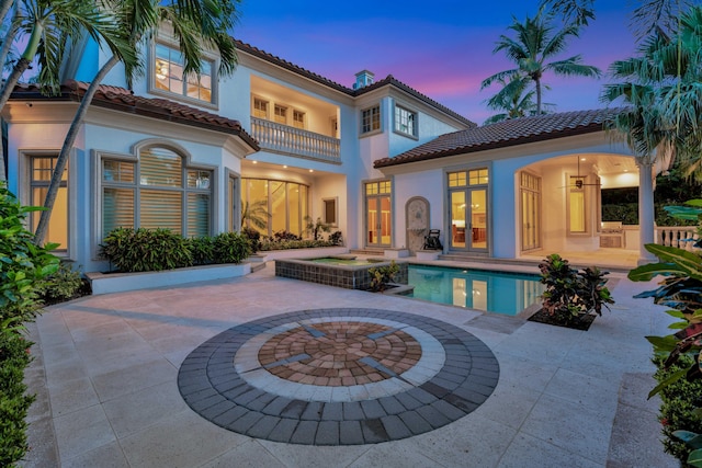 back of house at dusk featuring a balcony, a patio, stucco siding, and a tile roof