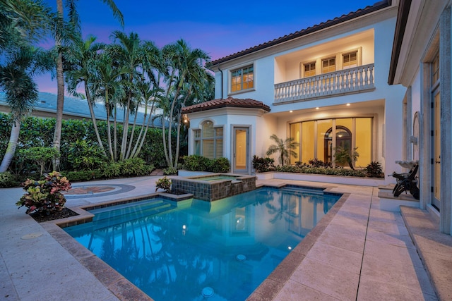 back of property at dusk featuring a pool with connected hot tub, stucco siding, a tiled roof, and a balcony