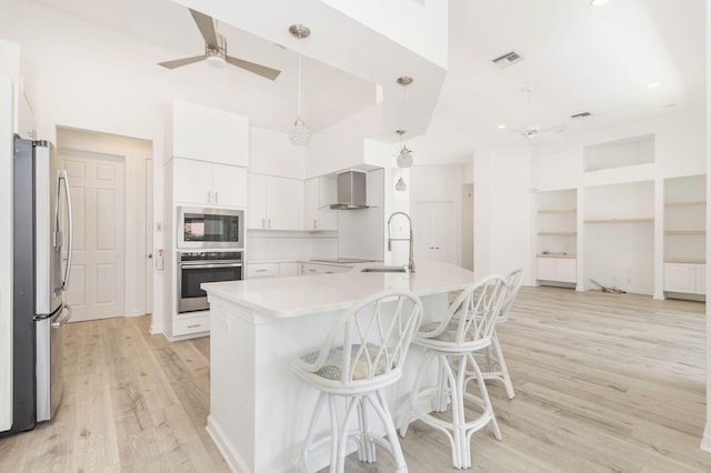 kitchen featuring wall chimney range hood, stainless steel appliances, sink, decorative light fixtures, and light wood-type flooring