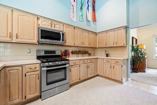 kitchen with light brown cabinetry, appliances with stainless steel finishes, decorative backsplash, light colored carpet, and a towering ceiling