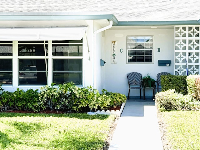 view of exterior entry featuring a shingled roof, a lawn, and stucco siding