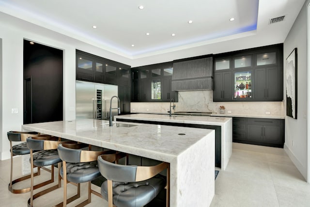 kitchen featuring a spacious island, backsplash, sink, light stone countertops, and stainless steel built in refrigerator