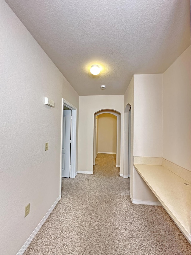 corridor featuring baseboards, a textured wall, arched walkways, a textured ceiling, and light colored carpet