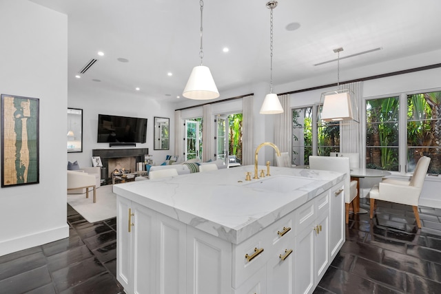 kitchen with light stone counters, white cabinetry, hanging light fixtures, and a kitchen island with sink