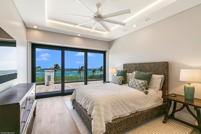 bedroom featuring light hardwood / wood-style floors, a water view, a tray ceiling, access to exterior, and ceiling fan