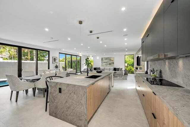 kitchen featuring a center island with sink, pendant lighting, sink, black electric cooktop, and light stone counters