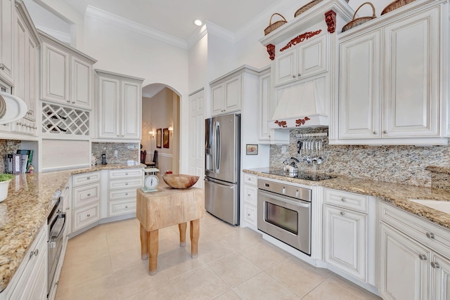 kitchen with appliances with stainless steel finishes, custom range hood, backsplash, ornamental molding, and light tile patterned floors