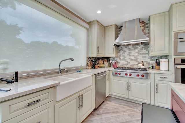 kitchen with wall chimney range hood, light wood-type flooring, backsplash, cream cabinetry, and stainless steel appliances