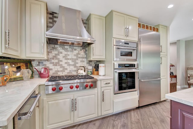 kitchen with stainless steel appliances, tasteful backsplash, cream cabinetry, light wood-type flooring, and wall chimney range hood