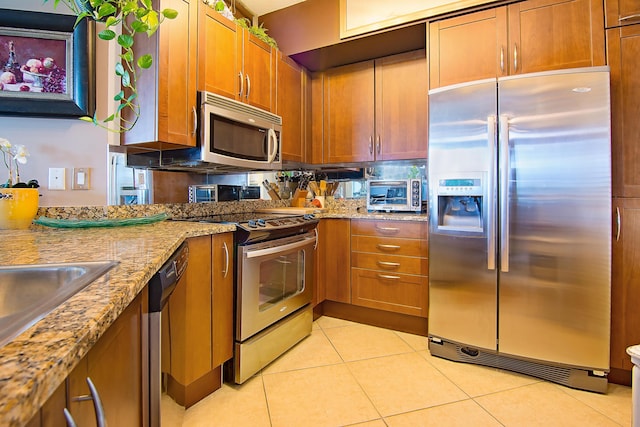 kitchen with a toaster, brown cabinets, appliances with stainless steel finishes, light tile patterned flooring, and light stone countertops