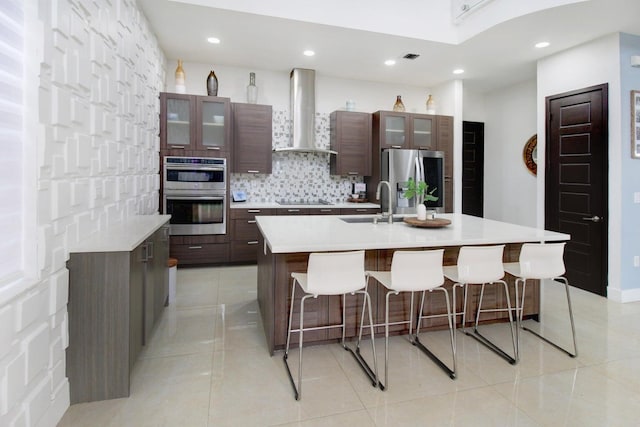 kitchen featuring appliances with stainless steel finishes, decorative backsplash, a center island with sink, wall chimney range hood, and light tile patterned flooring