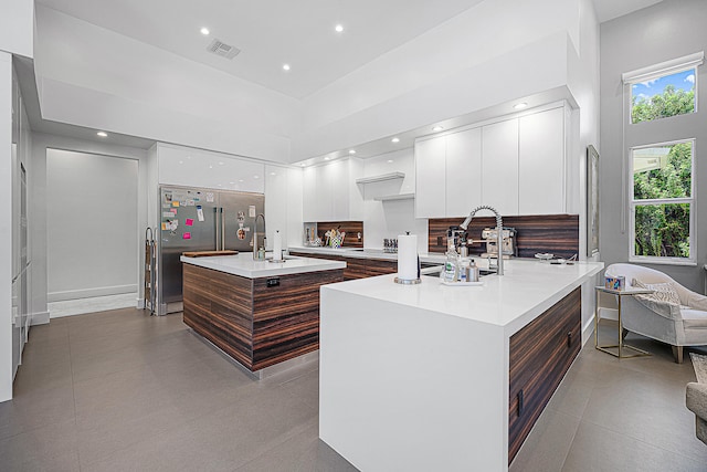 kitchen featuring stainless steel built in fridge, dark brown cabinetry, white cabinetry, a towering ceiling, and a center island with sink