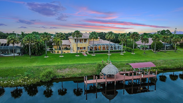 back of house at dusk with a lanai, a water view, a lawn, and boat lift