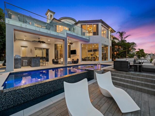 pool at dusk with ceiling fan, an outdoor kitchen, and outdoor lounge area