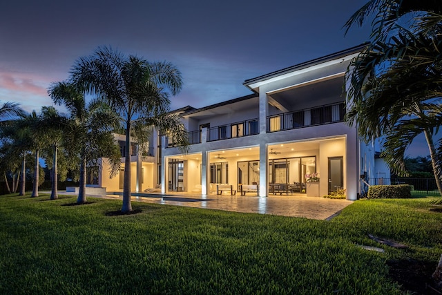 back house at dusk with a balcony, a patio area, and a lawn