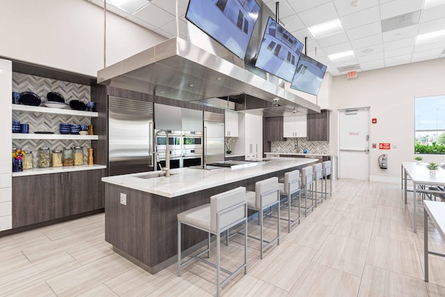 kitchen with built in fridge, light stone counters, an island with sink, and a drop ceiling