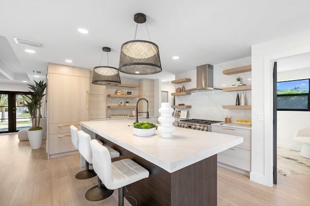 kitchen with light hardwood / wood-style floors, wall chimney exhaust hood, stainless steel range oven, decorative light fixtures, and a center island with sink