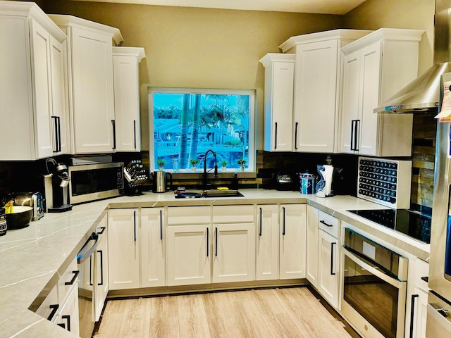 kitchen featuring appliances with stainless steel finishes, white cabinetry, wall chimney range hood, sink, and light wood-type flooring