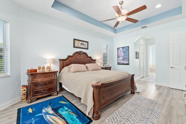 bedroom with ceiling fan, connected bathroom, light hardwood / wood-style flooring, and a tray ceiling