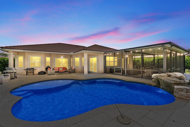 pool at dusk with a patio area, grilling area, outdoor lounge area, pool water feature, and a sunroom