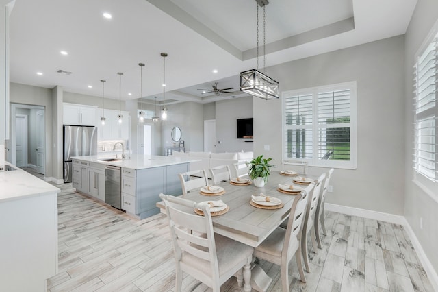 dining room with a raised ceiling, ceiling fan, sink, and light wood-type flooring