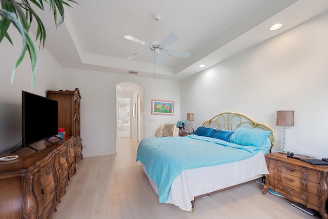 bedroom with ceiling fan, a raised ceiling, ensuite bathroom, and light wood-type flooring