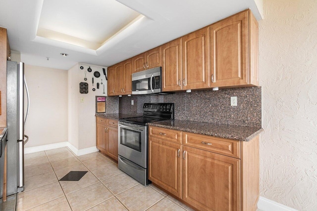 kitchen with light tile patterned floors, appliances with stainless steel finishes, a raised ceiling, backsplash, and dark stone counters